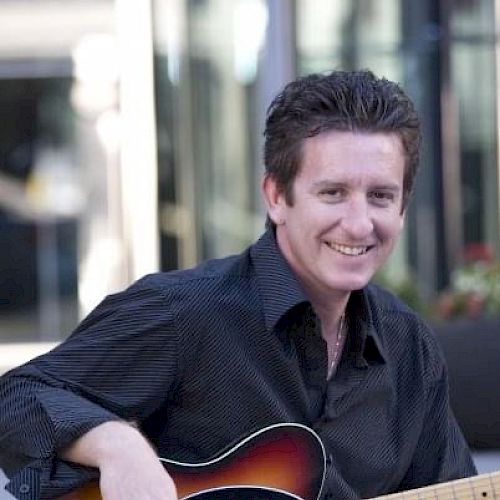 A person is sitting outdoors, smiling, and holding an electric guitar while wearing a black shirt and blue jeans.