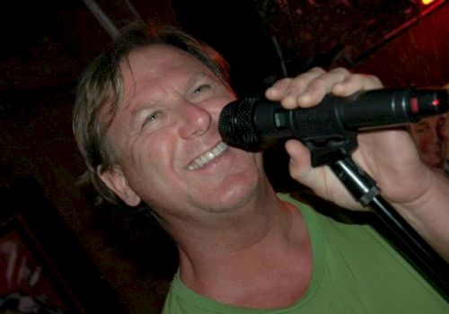 A person is smiling and singing into a microphone, wearing a green shirt, in a dimly lit setting.