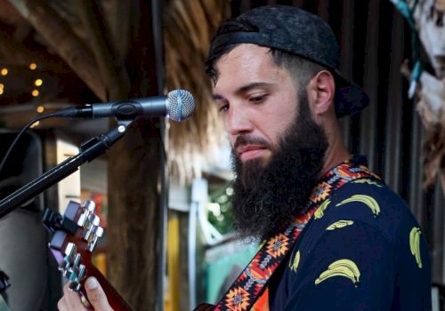 A bearded man wearing a cap and a banana-patterned shirt plays a guitar near a microphone stand, performing in an outdoor setting.