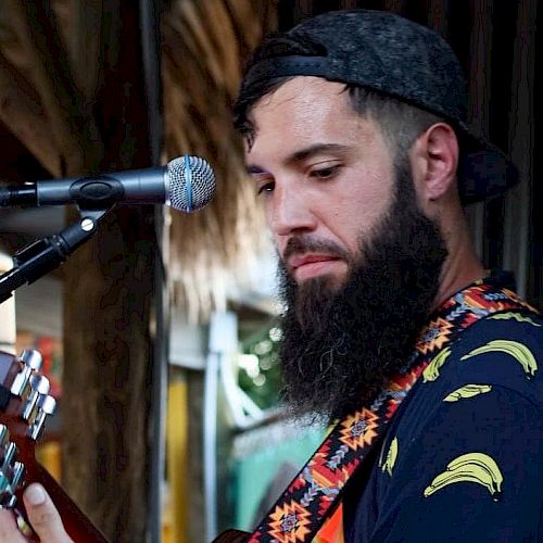 A bearded man wearing a cap and a banana-patterned shirt plays a guitar near a microphone stand, performing in an outdoor setting.