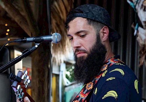 A man with a beard and a backward cap is playing a guitar and standing in front of a microphone, wearing a shirt with banana prints.