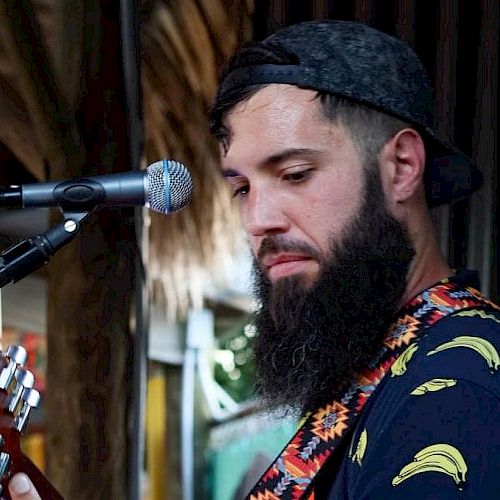 A man with a beard and a backward cap is playing a guitar and standing in front of a microphone, wearing a shirt with banana prints.