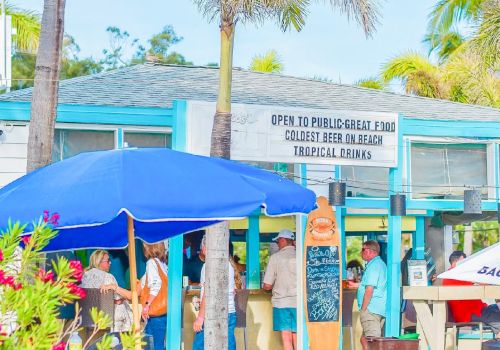 A small, tropical-themed cafe with palm trees, a blue umbrella, and a sign offering food, drinks, and tropical ambiance, with people gathered outside.