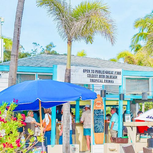 A small, tropical-themed cafe with palm trees, a blue umbrella, and a sign offering food, drinks, and tropical ambiance, with people gathered outside.