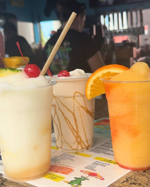 Three different frozen drinks with straws on a menu: a pina colada with cherries and pineapple, a caramel swirl drink, and a citrus drink with an orange slice.