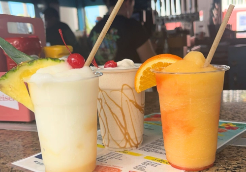 Three different frozen drinks with straws on a menu: a pina colada with cherries and pineapple, a caramel swirl drink, and a citrus drink with an orange slice.