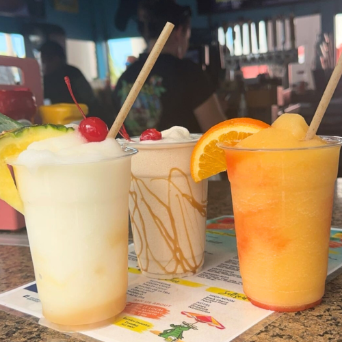 Three different frozen drinks with straws on a menu: a pina colada with cherries and pineapple, a caramel swirl drink, and a citrus drink with an orange slice.