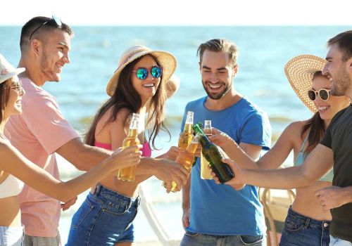 A group of friends on the beach is smiling and clinking their beer bottles together, celebrating a fun day in the sun.