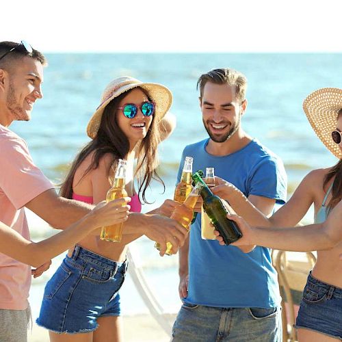 A group of friends on the beach is smiling and clinking their beer bottles together, celebrating a fun day in the sun.