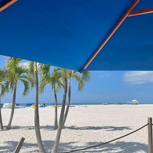 A tranquil beach scene with palm trees, white sand, and blue sky under a shaded umbrella, overlooking the ocean in the distance.