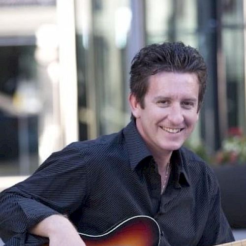 A person is sitting outdoors, holding a guitar and smiling at the camera. They are wearing a black shirt and blue jeans.