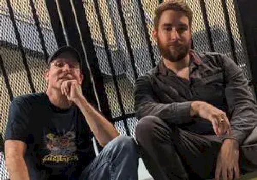 Two men are sitting on a step in front of a metal fence, one is wearing a black t-shirt and cap, the other is in a dark long-sleeve shirt.