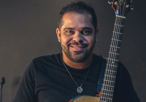 A person smiling while holding an acoustic guitar, wearing a dark long-sleeve shirt and a necklace, standing against a dark background.