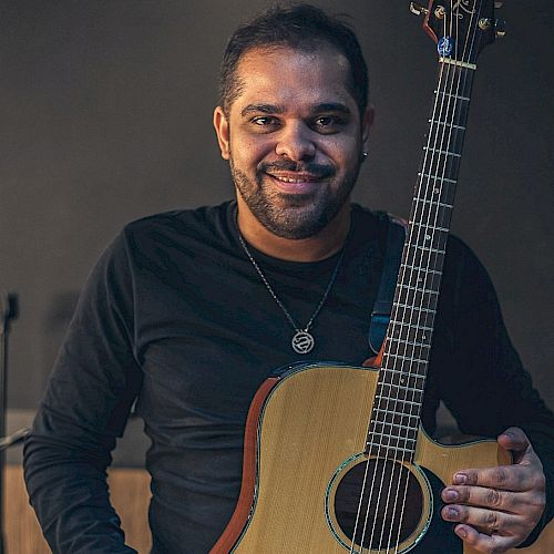 A person smiling while holding an acoustic guitar, wearing a dark long-sleeve shirt and a necklace, standing against a dark background.