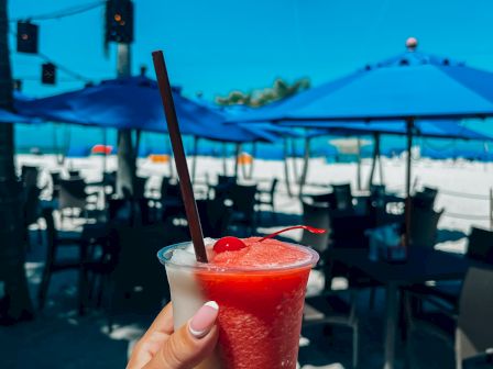 A person holds a tropical drink on a beach, with blue umbrellas and dining tables in the background. Palm trees and bright blue sky can be seen.