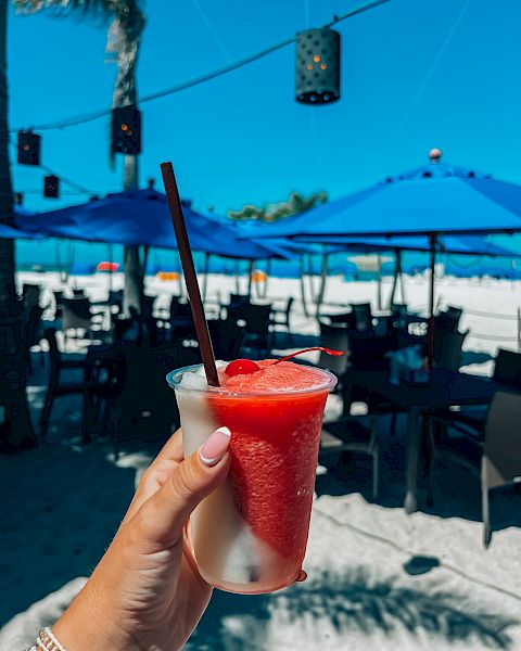 A hand holds a refreshing drink with a straw on a beach. Blue umbrellas and chairs are seen in the background under a bright blue sky.