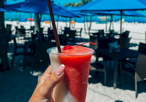 A hand holds a refreshing drink with a straw on a beach. Blue umbrellas and chairs are seen in the background under a bright blue sky.