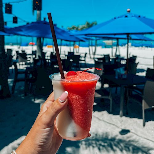 A hand holds a refreshing drink with a straw on a beach. Blue umbrellas and chairs are seen in the background under a bright blue sky.