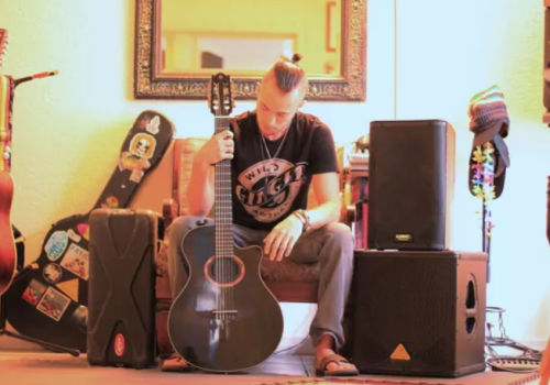 A person sits with a guitar in a room with musical equipment, including speakers, cases, and other guitars.