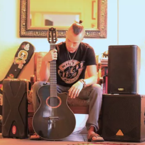 A person sits with a guitar in a room with musical equipment, including speakers, cases, and other guitars.