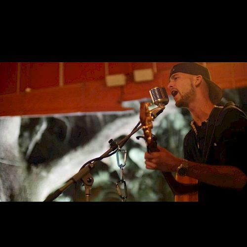 A man wearing a backwards cap is singing into a vintage microphone while playing an acoustic guitar in a dimly lit room.