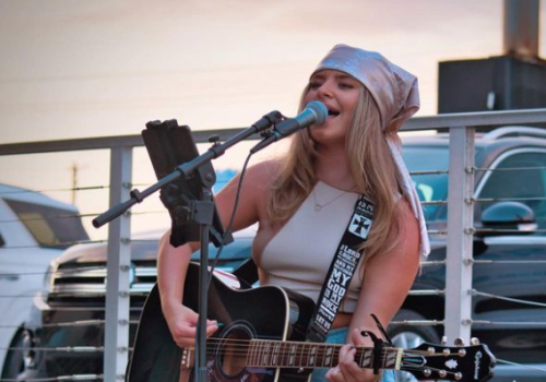 A woman with a guitar sings into a microphone outdoors, wearing a white bandana, tank top, shorts, and white boots. A guitar case and drink are nearby.