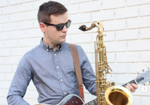 A person wearing sunglasses plays an acoustic guitar while a saxophone is attached to the guitar in front of a white brick wall.