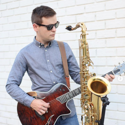 A person wearing sunglasses plays an acoustic guitar while a saxophone is attached to the guitar in front of a white brick wall.