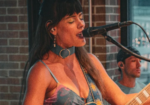 A woman is singing into a microphone and playing an electric guitar, with a drummer in the background, next to a brick wall and a window.