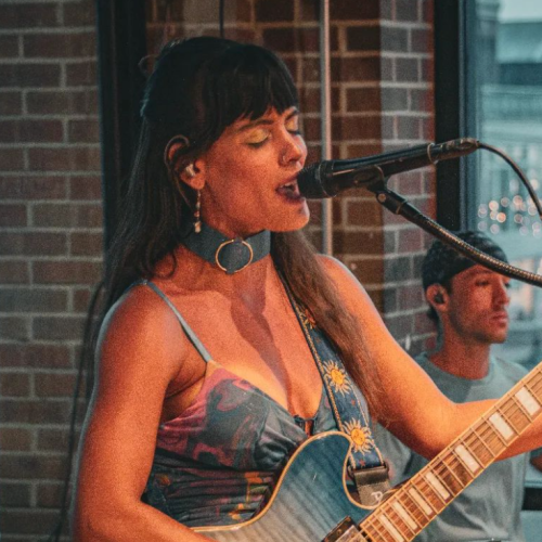 A woman is singing into a microphone and playing an electric guitar, with a drummer in the background, next to a brick wall and a window.