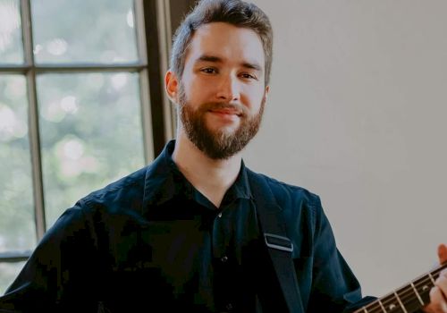 A person with a beard is smiling and playing an acoustic guitar near a window while wearing a black shirt.