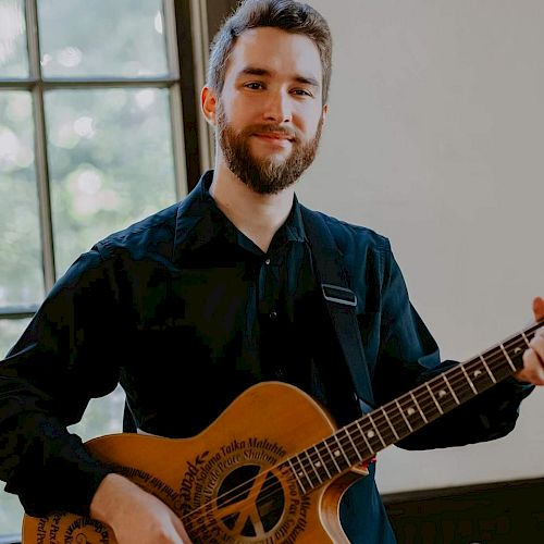 A person with a beard is smiling and playing an acoustic guitar near a window while wearing a black shirt.