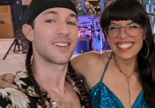 Two smiling people are posing for a selfie, with the man wearing a patterned shirt and the woman in a sparkly blue top and glasses, indoors.