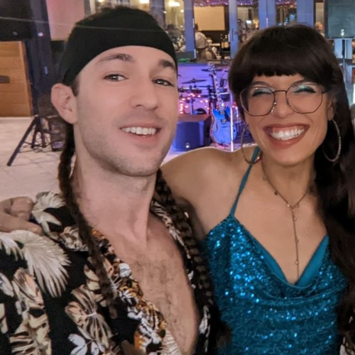 Two smiling people are posing for a selfie, with the man wearing a patterned shirt and the woman in a sparkly blue top and glasses, indoors.