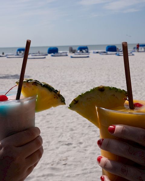 Two hands holding tropical drinks with pineapple garnishes on a sunny beach with umbrellas and lounge chairs in the background.