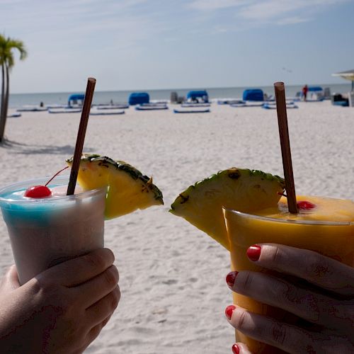 Two hands holding tropical drinks with pineapple garnishes on a sunny beach with umbrellas and lounge chairs in the background.