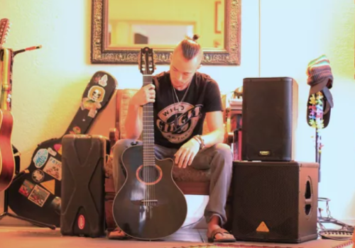 A person sits with a guitar amidst various musical equipment including speakers, cases, and another guitar standing behind them ending the sentence.