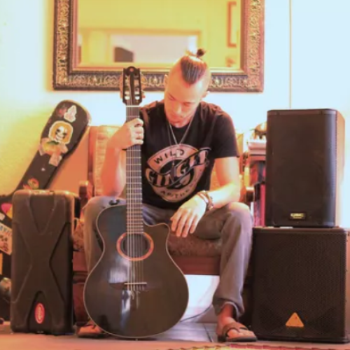 A person sits with a guitar amidst various musical equipment including speakers, cases, and another guitar standing behind them ending the sentence.