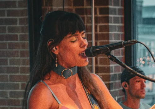 A woman is singing into a microphone while playing the guitar, with a person on the drums in the background, against a brick wall.