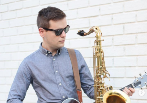 A person wearing sunglasses is playing a guitar while also holding a saxophone in front of a white brick wall.