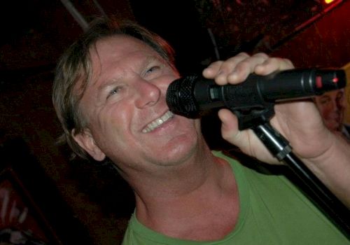 A person smiling and singing into a microphone while wearing a green shirt in a dimly lit setting.