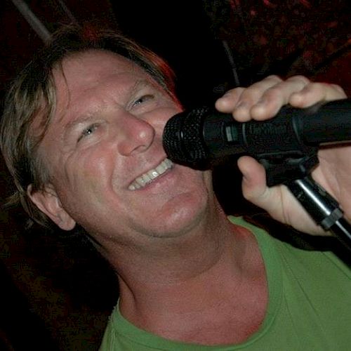 A person smiling and singing into a microphone while wearing a green shirt in a dimly lit setting.