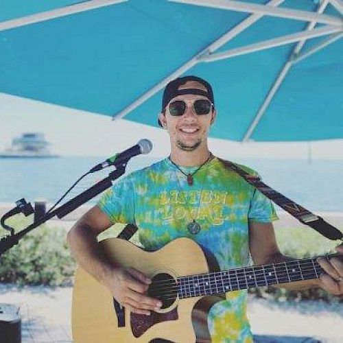 A man is smiling under a blue umbrella by the water while holding a guitar, standing in front of a microphone on a sunny day, wearing sunglasses.