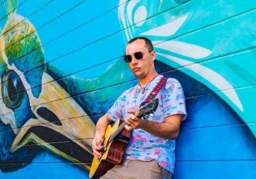 A man in sunglasses and casual attire is playing a guitar against a brightly colored mural wall, featuring a large bird graphic.