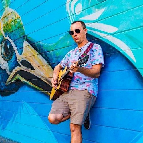 A man in sunglasses and casual attire is playing a guitar against a brightly colored mural wall, featuring a large bird graphic.