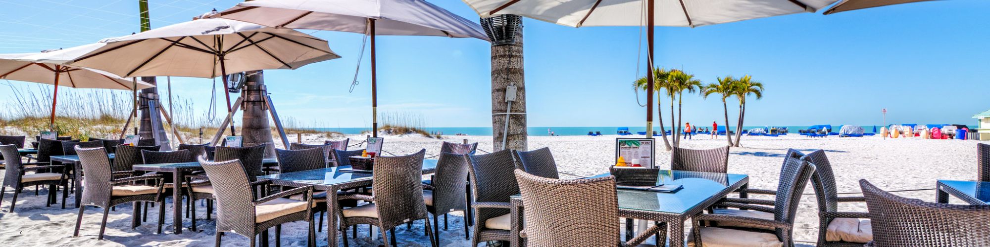 Outdoor restaurant with tables and chairs on a sandy beach, shaded by umbrellas, surrounded by palm trees under a clear blue sky.