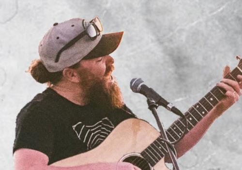 The image shows a bearded person wearing a cap and sunglasses, playing an acoustic guitar and singing into a microphone.