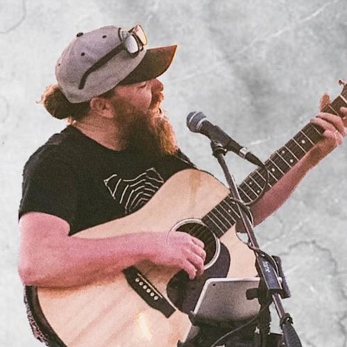 The image shows a bearded person wearing a cap and sunglasses, playing an acoustic guitar and singing into a microphone.