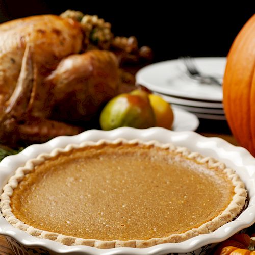 A Thanksgiving meal setup featuring a roast turkey, a pumpkin pie, pumpkins, and fruits, with plates and utensils on the table.