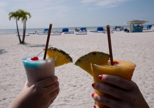 Two hands holding tropical drinks with fruit garnishes on a sandy beach, overlooking the ocean and beach chairs under umbrellas.